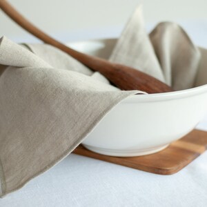 May include: A white ceramic bowl with a wooden spoon and a linen napkin inside. The bowl is sitting on a wooden cutting board.