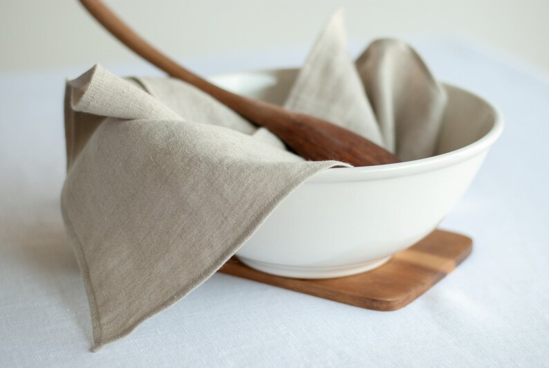 May include: A white ceramic bowl with a wooden spoon and a linen napkin inside. The bowl is sitting on a wooden cutting board.