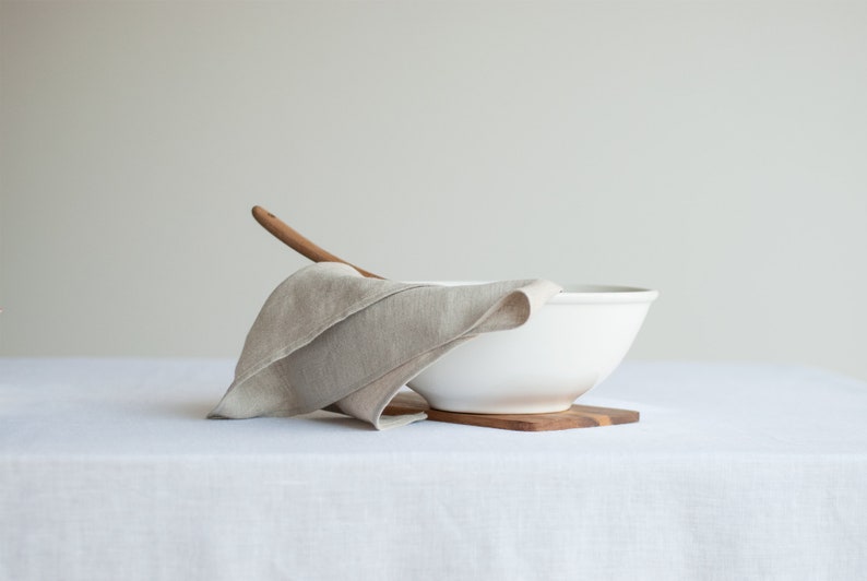 May include: A white ceramic bowl with a wooden spoon resting on a linen napkin. The bowl is sitting on a wooden coaster on a white linen tablecloth.