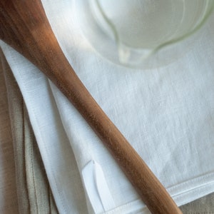 May include: A wooden spoon lying on top of two folded linen tea towels, one white and one beige.