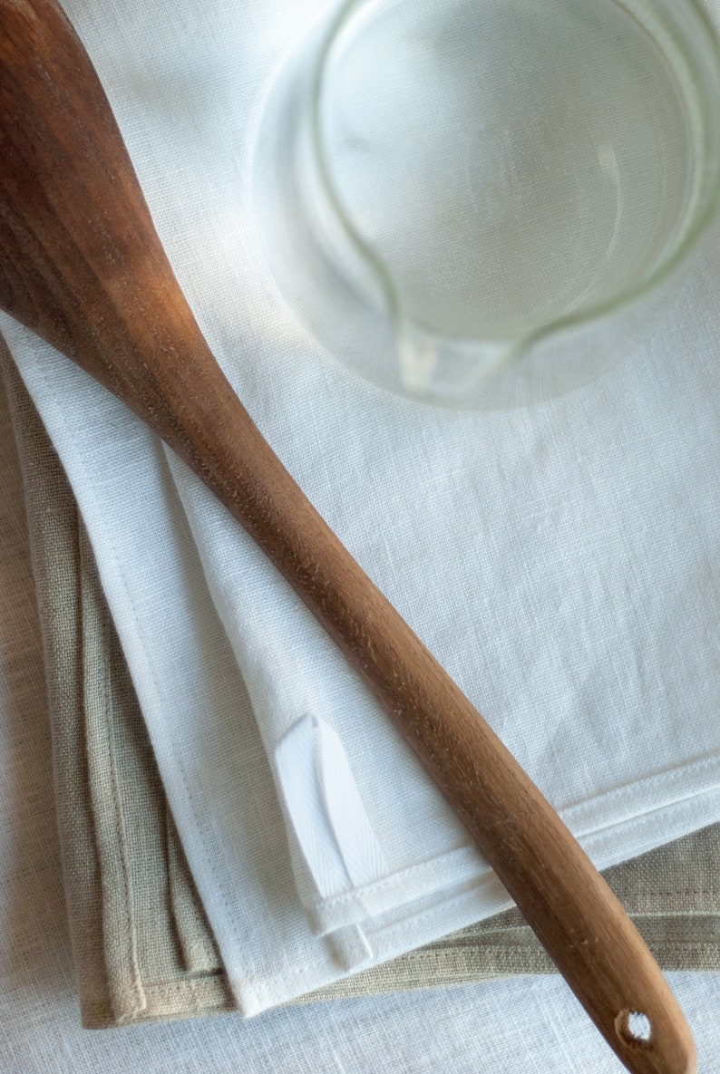 May include: A wooden spoon lying on top of two folded linen tea towels, one white and one beige.