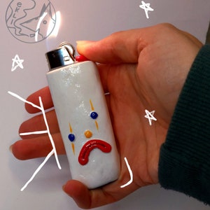 Può includere: Accendino bianco con un viso triste dipinto con dettagli gialli, rossi e blu. L'accendino è tenuto in una mano.