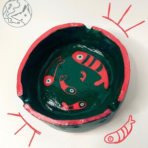 Può includere: Un posacenere in ceramica verde con un bordo rosa. L'interno del posacenere è dipinto con pesci rossi e bianchi.