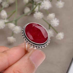 Può includere: Un anello d'argento con una grande pietra di rubino ovale. Il rubino è di un rosso intenso ed è incastonato in una fascia d'argento decorativa con un bordo a forma di corda. L'anello è tenuto da una mano.