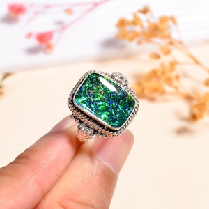 Puede incluir: Un anillo de plata con una piedra preciosa verde iridiscente rectangular. La piedra preciosa tiene un aspecto texturizado con tonos verdes y azules. El anillo tiene un engaste de plata detallado y ornamentado con un borde trenzado y detalles decorativos.