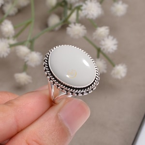 May include: Oval-shaped ring with a light green stone set in a silver band. The stone is surrounded by a twisted rope design. The ring is held up by a hand.