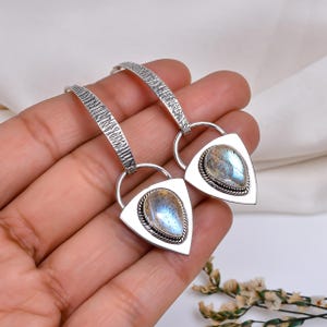 Puede incluir: Pendientes de plata con una parte superior curvada y texturizada y un colgante triangular. Cada colgante presenta una piedra preciosa iridiscente en forma de lágrima con tonos azules y grises, enmarcada por un borde plateado. Fondo blanco.