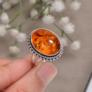 Può includere: Anello ovale con pietra preziosa in ambra con fascia in argento. La pietra d'ambra è di colore arancione caldo ed è incastonata in una montatura d'argento decorativa. L'anello è tenuto da una mano, che mostra il gioiello.