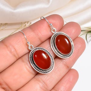 May include: Oval-shaped carnelian earrings with a silver-colored rope-like border. The earrings have a polished, reddish-orange stone and a simple hook closure. The earrings are displayed against a soft, neutral background.