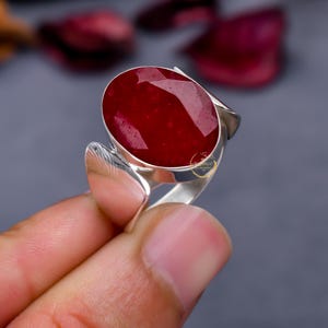 May include: A silver ring featuring a large, oval-cut, deep red gemstone. The ring has a modern design with a smooth, polished band and decorative side accents. The gemstone is the focal point, showcasing its rich color and facets.