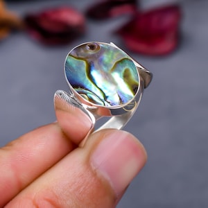 Puede incluir: Un anillo de plata con una incrustación ovalada de concha de abulón. La concha tiene colores iridiscentes verdes, azules y morados.