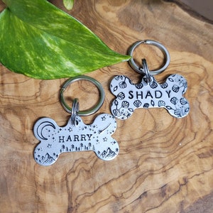 May include: Two silver dog tags shaped like a bone. The top tag features a seashell design and the text "SHADY". The bottom tag has a mountain and forest design with the text "HARRY".