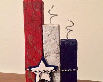 May include: A patriotic wooden decoration featuring three rectangular blocks painted red, white, and blue. Each block has a silver wire spiral at the top. A star with a blue border and white center is tied to the blocks with black and white twine.