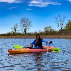 May include: A woman in a blue shirt and life vest sits in a bright orange kayak on a calm lake. She is holding a green paddle and pouring water from a blue container.