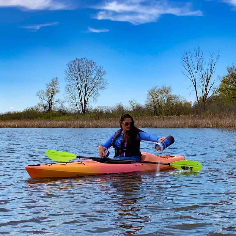 May include: A woman in a blue shirt and life vest sits in a bright orange kayak on a calm lake. She is holding a green paddle and pouring water from a blue container.