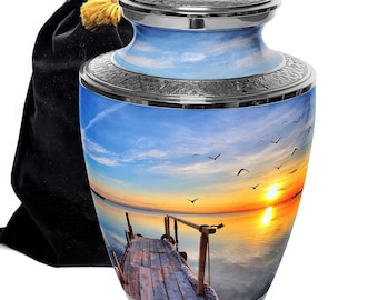 May include: A silver urn with a sunset scene featuring a wooden dock extending into a body of water. The urn has a silver lid and a black velvet bag.