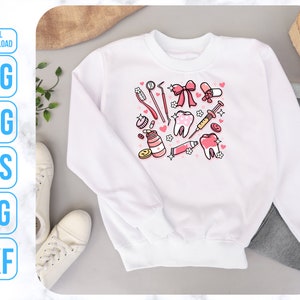 Op de afbeelding: Wit sweatshirt met een roze en rood ontwerp met verschillende tandheelkundige instrumenten, waaronder tandenborstels, floss en een spuit. Het ontwerp bevat ook harten en tanden.