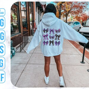 May include: A white hoodie with a design of purple, black, and gold bows on the back. The hoodie is being worn by a person standing on a sidewalk.