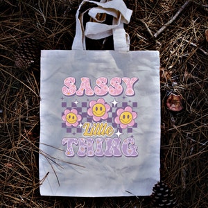 May include: A beige tote bag with the words "SASSY LITTLE THING" in a retro font. The design includes pink flowers, smiley faces, and a checkered pattern in purple and white. The bag has two handles.