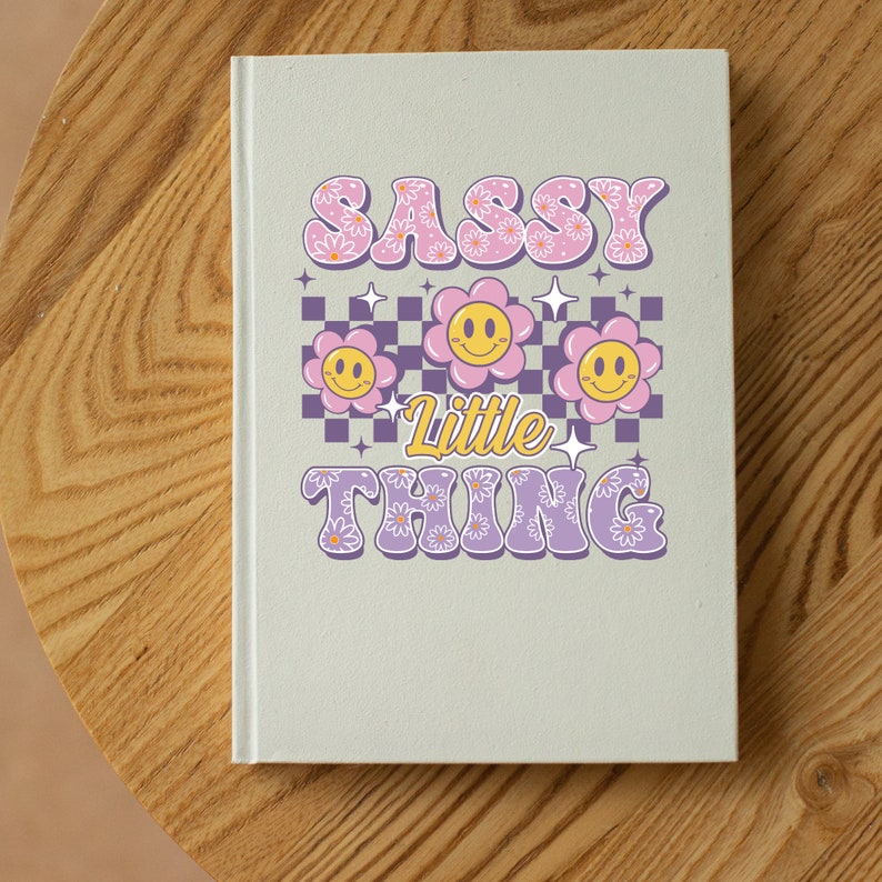 May include: A light green notebook with the words "SASSY Little THING" in a retro font. The design includes pink daisy flowers, smiley face flowers, and a purple and white checkered pattern.