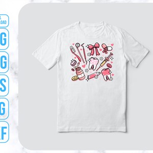 Op de afbeelding: Een wit T-shirt met een roze en wit ontwerp van tandheelkundige instrumenten, een tand en een strik. Het ontwerp bevat een tandenborstel, tandpasta, floss, een spuit en een hart.