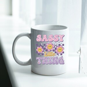 May include: White ceramic mug with the text "SASSY Little THING" in a retro font. The design includes smiling flower graphics in pink, yellow, and purple, set against a checkered background. The mug has a curved handle.