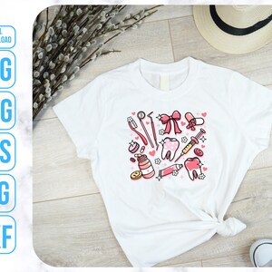 Op de afbeelding: Wit T-shirt met een roze en wit ontwerp van tandheelkundige instrumenten, waaronder een tandenborstel, tandpasta, floss en een spuit. Het ontwerp bevat ook een roze strik en harten.