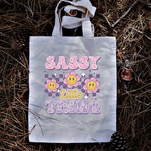 May include: A beige tote bag with the words "SASSY Little THING" in a retro font. The design includes smiling flower graphics and a checkered pattern in pink, purple, and yellow. The bag has two handles.