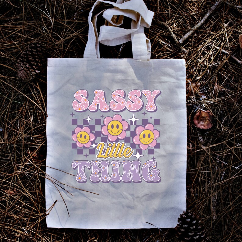 May include: A beige tote bag with the words "SASSY Little THING" in a retro font. The design includes smiling flower graphics and a checkered pattern in pink, purple, and yellow. The bag has two handles.