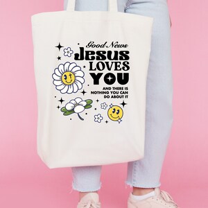 May include: White canvas tote bag with a black and white graphic design featuring the text "Good News Jesus Loves You" and a daisy with a smiley face. The text below the graphic says "And there is nothing you can do about it."