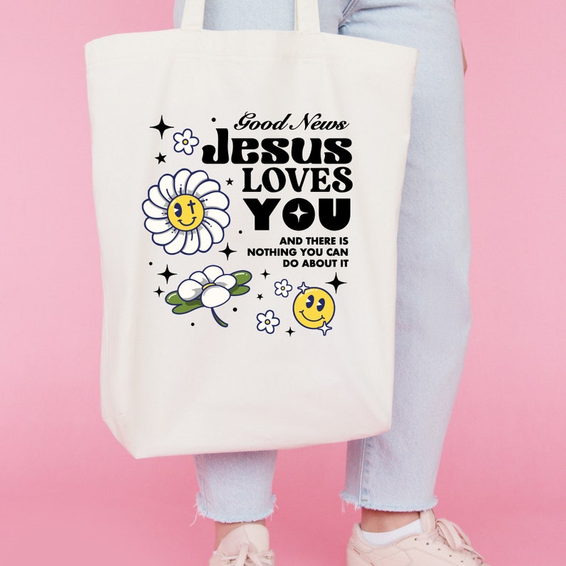 May include: White canvas tote bag with a black and white graphic design featuring the text "Good News Jesus Loves You" and a daisy with a smiley face. The text below the graphic says "And there is nothing you can do about it."