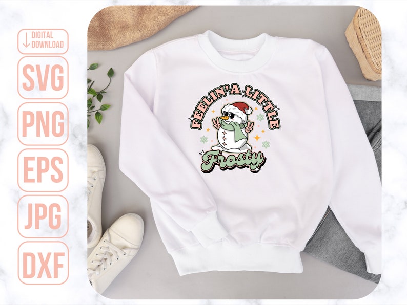 Może przedstawiać: Biała bluza z nadrukiem przedstawiającym bałwana w czapce Mikołaja i okularach przeciwsłonecznych. Tekst "Feelin' a little Frosty" jest napisany r&oacute;żowymi i zielonymi literami.
