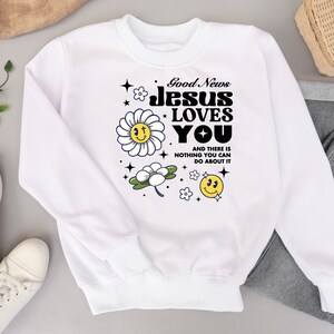 May include: White sweatshirt with a graphic design that says "Good News Jesus Loves You" with a daisy and smiley face design.