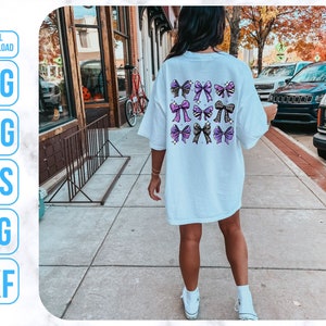May include: A white t-shirt with a design of purple and black bows on the back. The shirt is being worn by a person walking down a sidewalk.