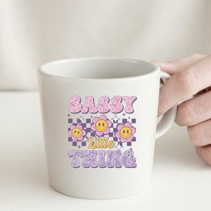 May include: White ceramic mug with the text "SASSY Little THING" in pink and yellow. The design includes three smiling flower graphics on a purple and white checkered background. The mug is held by a hand.