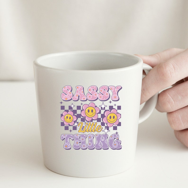 May include: White ceramic mug with the text "SASSY Little THING" in pink and yellow. The design includes three smiling flower graphics on a purple and white checkered background. The mug is held by a hand.