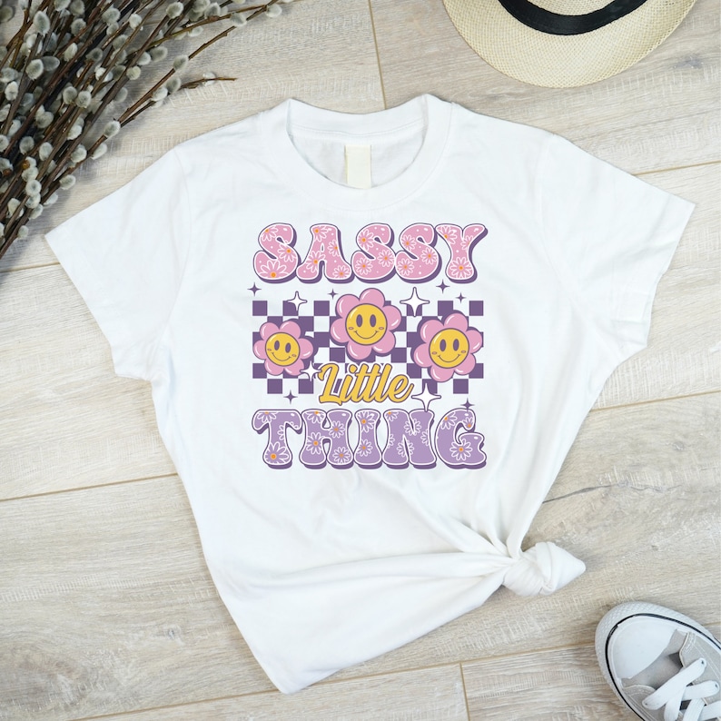 May include: White t-shirt with the text "SASSY Little THING" in a retro font. The design includes smiling flower graphics and a checkered background in pink, purple, and yellow.