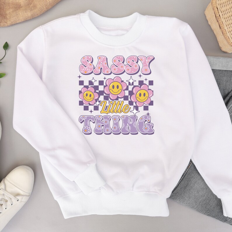 May include: White sweatshirt with the text "SASSY Little THING" in a retro font. The design includes smiling flower graphics in pink and yellow, set against a checkered and star background. A casual, comfortable top.