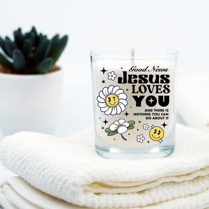 May include: A white candle with a glass jar and a black and white graphic design featuring the text "Good News Jesus Loves You" and a daisy with a smiley face. The text below the graphic says "And there is nothing you can do about it."
