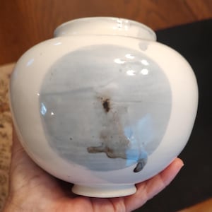 May include: A white ceramic vase with a rounded body and a small opening. The vase features a large, light blue and gray abstract design on the front. The vase is held in a person's hand.