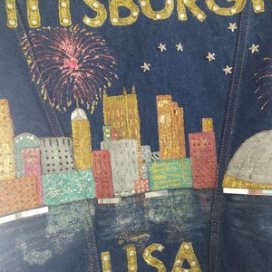 May include: A denim jacket with a glittery design of the Pittsburgh skyline, fireworks, and the word "PITTSBURGH" in gold letters. The jacket also has the word "LISA" in gold letters at the bottom.