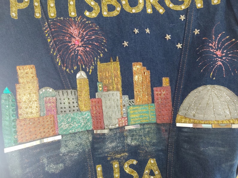 May include: A denim jacket with a glittery design of the Pittsburgh skyline, fireworks, and the word "PITTSBURGH" in gold letters. The jacket also has the word "LISA" in gold letters at the bottom.