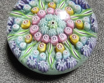 May include: A round glass paperweight with a floral design. The center features a cluster of light blue and pink elements, surrounded by a ring of pink and yellow flower-like shapes. Green leaves and blue floral accents complete the design.