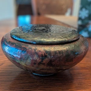 May include: A handcrafted, ceramic lidded bowl with a textured, dark metallic finish. The bowl's body features a blend of brown, red, and black hues, creating a unique, rustic appearance. The lid has a hammered texture, adding to its artisanal charm.