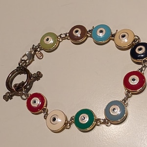 May include: A colorful bracelet featuring a series of circular charms in various colors, including red, green, blue, and cream. Each charm has a central black dot and is outlined in gold. The bracelet is connected by a delicate chain and has a toggle clasp.