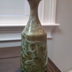 May include: A small, green and white ceramic vase with a unique, speckled pattern. The vase has a narrow neck and a wide base.