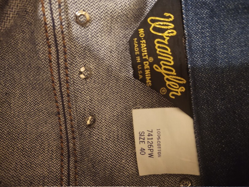 May include: A blue denim Wrangler label with the text "Wrangler No-Fault Denims Made in U.S.A." and a white label with the text "100% Cotton 74126PW SIZE 40".