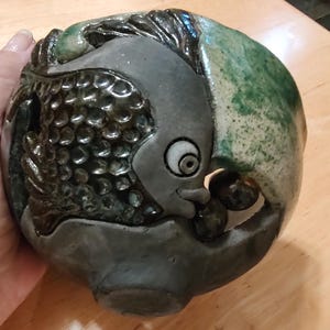 May include: A ceramic candle holder with a fish design. The candle holder is a rounded shape with a gray fish design on the front and green accents. The fish has a smiling face and a textured body. The candle holder is on a wooden surface.