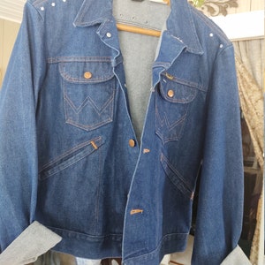 May include: A blue denim jacket with a brown leather label that says "Wrangler" and a row of silver studs on the shoulder. The jacket has a pointed collar, a button closure, and two chest pockets.