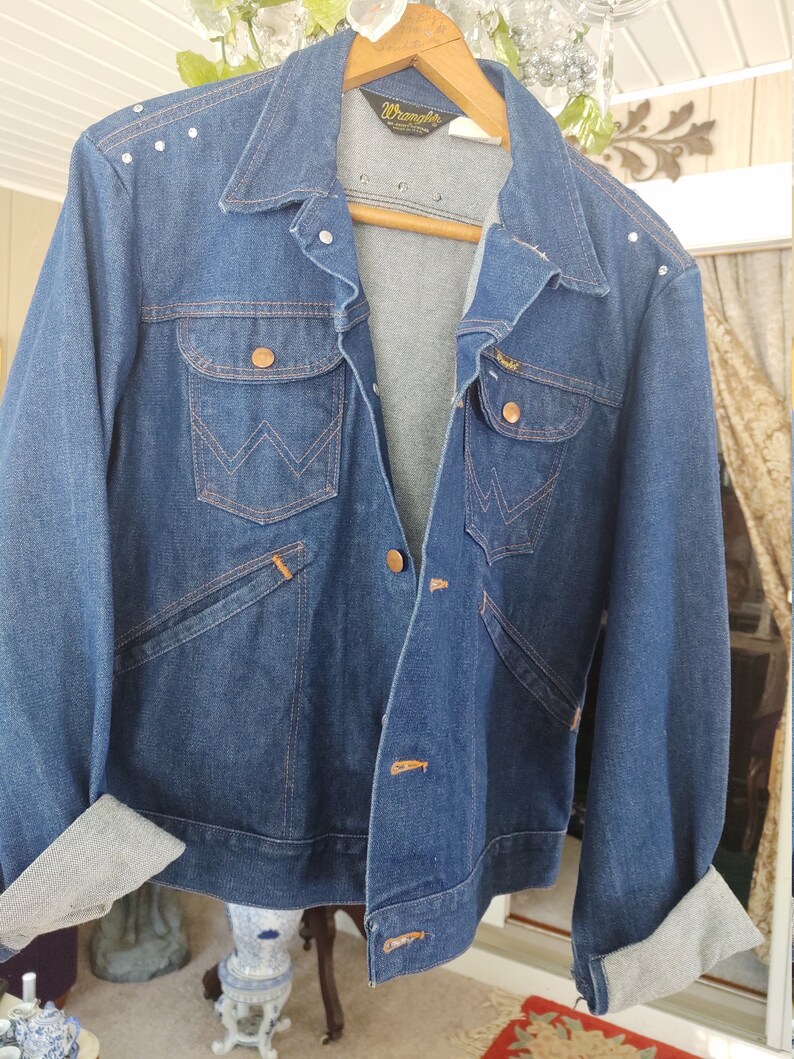 May include: A blue denim jacket with a brown leather label that says "Wrangler" and a row of silver studs on the shoulder. The jacket has a pointed collar, a button closure, and two chest pockets.